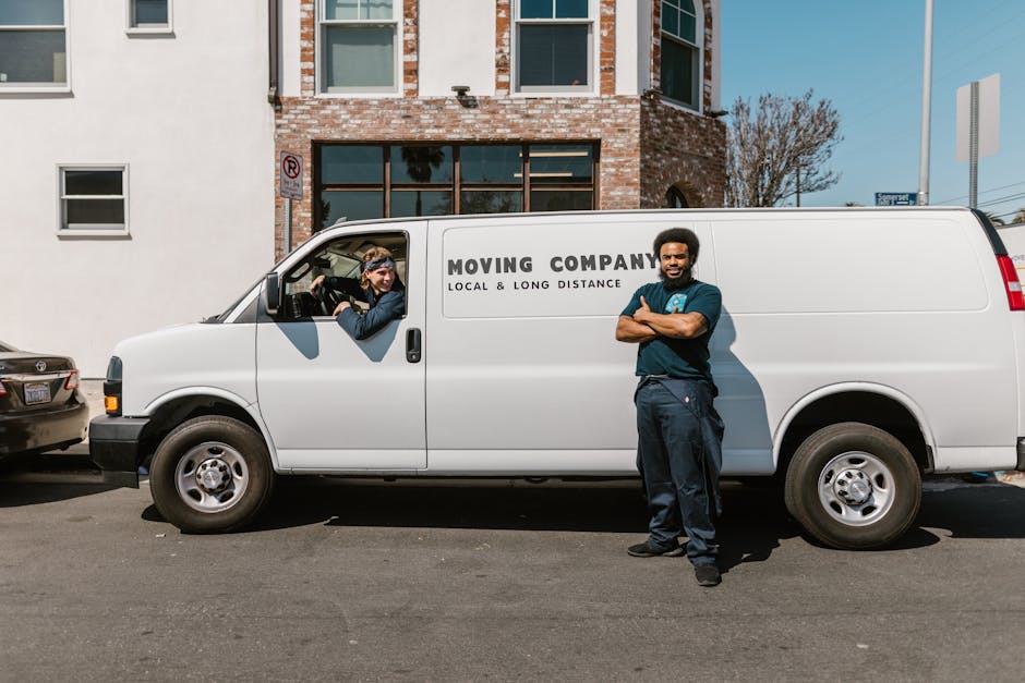 A man with dark skin and a beard, dressed in a navy blue uniform, stands with arms crossed outside a white moving van, which has the text 'MOVING COMPANY LOCAL & LONG DISTANCE' printed on its side. Th