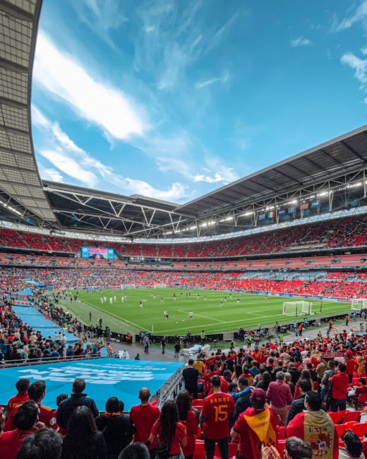 A wide-angle view of a packed football stadium during a match, with a clear blue sky and scattered clouds overhead. The stadium's multi-tiered seating is filled with spectators dressed in red and other colours, watching the players on the green pitch below. Visible elements include the goalposts, players actively engaged in the game, and a large digital scoreboard hanging above the stands. The floodlights are turned off as daylight illuminates the scene. The stadium structure features metal framing and a partially covered upper tier, with a road or pathway around the outer edge. This image exemplifies a high-capacity sports venue, which could be relevant to relocation or event planning services such as those offered by Man with Van Wembley, especially in contexts relating to logistical arrangements for large gatherings or transport of equipment and supplies.