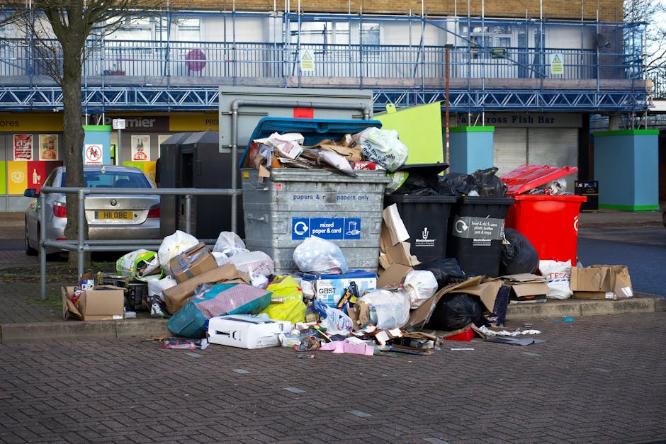 An overflowing waste disposal area outside a commercial building in Wembley, with multiple black, grey, and red rubbish bins filled to capacity, overflowing with cardboard boxes, plastic bags, paper, and household waste piled on the pavement. Some waste, including flattened cardboard boxes, paper, plastic wraps, and small packaged items, is scattered on the ground around the bins. The scene shows a mix of packaging materials and household refuse, with a background featuring retail shopfronts, a silver car parked nearby, and a tree on the sidewalk. This waste collection area reflects common household and commercial waste that often requires proper disposal or professional removal, such as that provided by companies like Man with Van Wembley, especially during home relocation or packing and moving processes approved by Brent Council's bulky waste rules.