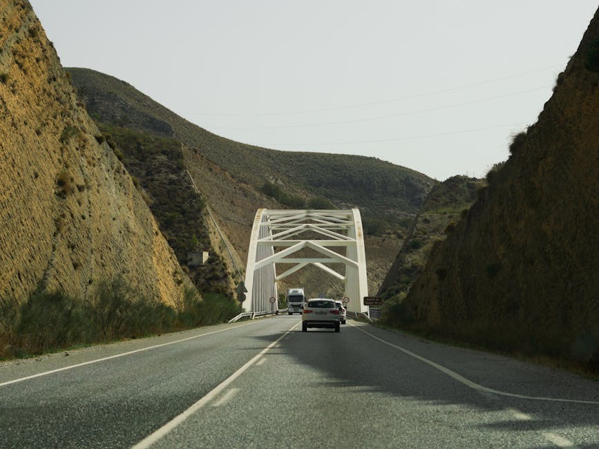 A two-lane road with white dashed centre lines passing through a narrow mountain pass, flanked by steep slopes covered in grass and sparse shrubbery. Several vehicles, including a white van, are traveling towards a white arched bridge spanning the road ahead. The bridge's structural framework is visible, with multiple support beams creating a geometric pattern against the sky. The environment appears to be rural with natural surroundings, and the lighting indicates daytime with clear weather conditions. This scene illustrates a challenging narrow access route that may require careful planning during home relocation or furniture transport, as managed by companies like Man with Van Wembley, particularly when navigating bridges and tight spaces.