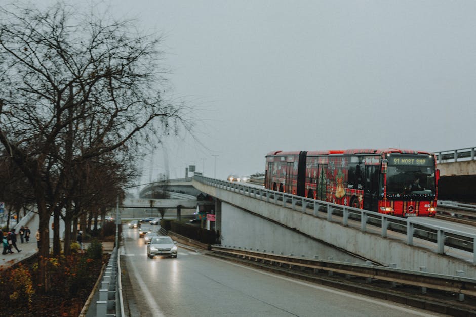 A red double-decker bus is positioned on the upper level of a narrow bridge with a metal railing, crossing over a busy street in Wembley. In the background, there are multiple vehicles including cars and vans traveling along the road below the bridge, with some appearing to be in motion due to the slight blur. The scene is set during daytime with overcast skies, creating diffused lighting. To the left, a few pedestrians are walking along a sidewalk lined with leafless trees, indicating a colder season. The bridge's wrought iron is visible beneath the bus, and the street below features a mix of parked and moving vehicles. This urban setting, featuring detailed transportation and street elements, relates to the context of house removals and moving logistics, emphasizing the challenges of navigating narrow or elevated access points as seen in home relocation strategies. Man with Van Wembley, a professional removals service, considers such logistical factors in planning efficient furniture transport and packing during a house move on Bridge Road.