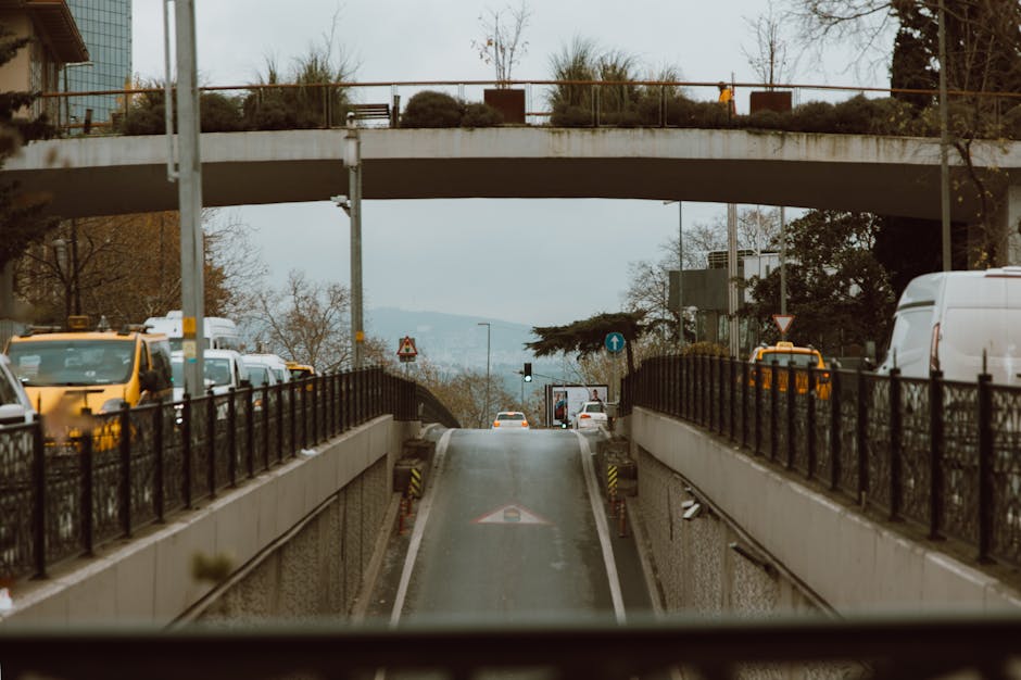 A two-lane road with white dashed centre lines passing through a narrow mountain pass, flanked by steep slopes covered in grass and sparse shrubbery. Several vehicles, including a white van, are traveling towards a white arched bridge spanning the road ahead. The bridge's structural framework is visible, with multiple support beams creating a geometric pattern against the sky. The environment appears to be rural with natural surroundings, and the lighting indicates daytime with clear weather conditions. This scene illustrates a challenging narrow access route that may require careful planning during home relocation or furniture transport, as managed by companies like Man with Van Wembley, particularly when navigating bridges and tight spaces.