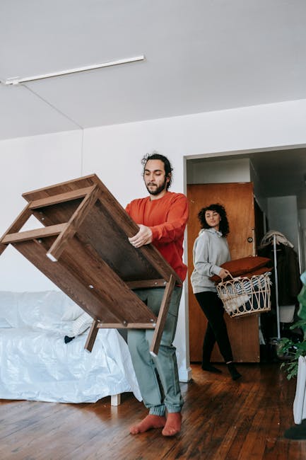 A man wearing a red long-sleeved shirt and grey pants is lifting a wooden dining table with a rectangular top and four legs inside a room, preparing to load it onto a vehicle as part of a home relocation. He is barefoot and standing on a polished wooden floor near a white sofa covered with a plastic sheet, indicating a careful packing process. In the background, a woman with curly dark hair, dressed in a grey hoodie and black pants, is carrying a wicker laundry basket filled with folded textiles toward the doorway, possibly to be loaded for transport. The room has white walls and a ceiling with a mounted ceiling light. To the right, a doorway leads to another area with visible wooden doors and what appears to be packing or moving equipment, suggesting a coordinated furniture transport operation. The scene depicts a household moving process with furniture disassembly, packing, and preparation for transportation arranged by Man with Van Wembley, highlighting the logistical aspects of a communal lift within an estate move on Empire Way in Wembley.