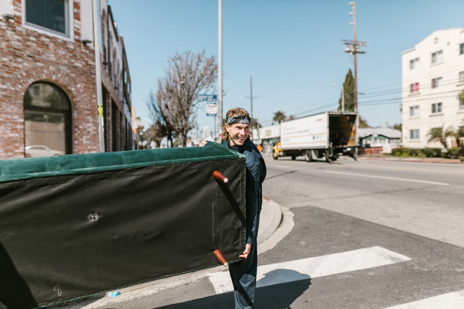 A young man with light brown hair, wearing a dark jacket and a sports headband, is outside on a clear day, carrying a large, black moving box with green fabric padding on top. He is smiling and appears to be preparing for a home relocation process as part of a furniture transport or packing and moving service. The scene is set at the corner of a street with a brick building with arched windows, a tree with bare branches, and a busy road with a white truck and other vehicles in the background. The pavement has a curb, and a pedestrian crossing is visible nearby. This image may be used to illustrate the logistical steps involved in a house removal in Wembley, managed by Man with Van Wembley, including loading boxes and furniture for an estate move on Empire Way, with emphasis on communal lift management and moving preparations.