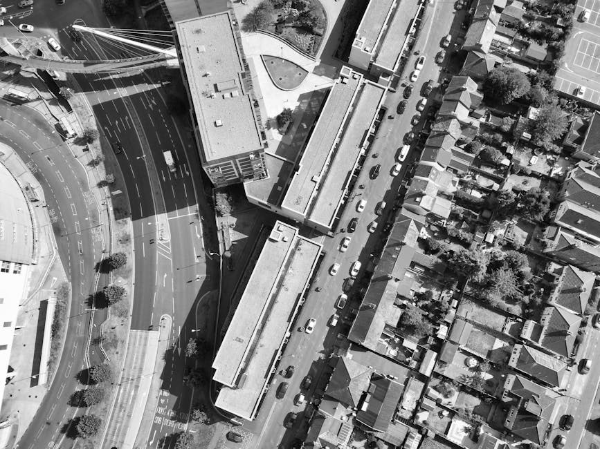 Aerial view of a street in Wembley showing parked cars along a narrow road next to residential houses with gardens, trees, and rooftops. On the left side of the image, there is a large building with a flat roof adjacent to a busy multi-lane road, which features lane markings, traffic lights, and trees lining the pavement. The residential area on the right side displays a row of terraced houses with pitched roofs, small front yards, and driveways. In the foreground, a household furniture removal process is visible, with people lifting and carrying cardboard boxes, plastic-wrapped furniture, and household appliances from a property onto a man with van from Wembley, which is parked nearby. The loading area includes a small mobile stair or trolley to facilitate moving items into the van, with blankets and straps used to secure items during transport as part of home relocation or furniture transport operations. Natural daylight illuminates the scene, highlighting the logistical movement involved in house removals in the Wembley area.