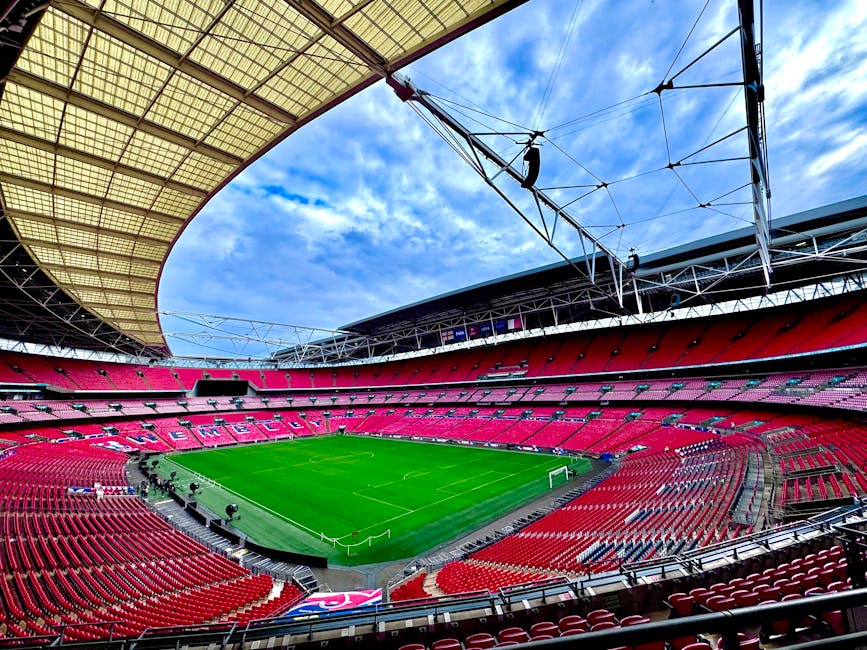 A panoramic view of a large football stadium filled with spectators, with the playing field visible in the center featuring players practicing and a goalpost on one side. The stadium has multiple tiers of seating packed with fans, some wearing team jerseys and colors, with red and white clothing prominent among the crowd. The sky above is partly cloudy with patches of blue, and the stadium's modern roof structure arches over part of the seating area. The image is taken from the perspective of an observer standing behind a section of seating, capturing the overall scene during daylight. Although the scene focuses on sports and spectator seating, the context relates to the moving process for house removals, emphasizing the importance of accessing and navigating large, busy venues during logistics operations, which Man with Van Wembley may coordinate during relocation projects.
