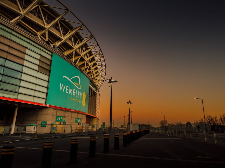 Exterior view of Wembley Stadium at sunset, with a large digital sign displaying the Wembley logo and the word 'CONNECTED' on the curved facade. The area in front of the stadium includes empty pavement, black bollards, and tall street lamps, indicating an outdoor space potentially used for home relocation or furniture transport operations. Inside, there are visible cardboard boxes, plastic wrapping, and fabric coverings stacked on a trolley near the entrance, suggesting packing and loading activities. The lighting conditions highlight the stadium's structural details and the quiet, organized environment, which is relevant to the logistics involved in house removals or relocation services provided by Man with Van Wembley. The setting implies a preparation or post-event clearance phase, emphasizing the importance of efficient packing and transport during home moves or furniture transport processes.
