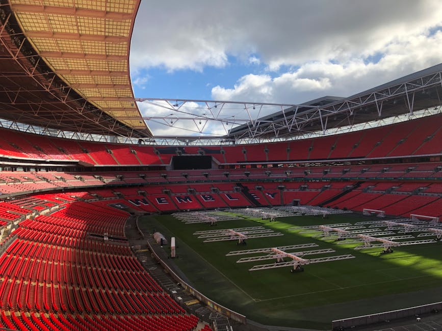 Inside a large, modern football stadium with a well-maintained green pitch at the center, surrounded by multiple tiers of red seating areas. The roof structure is partially visible, featuring a beige,