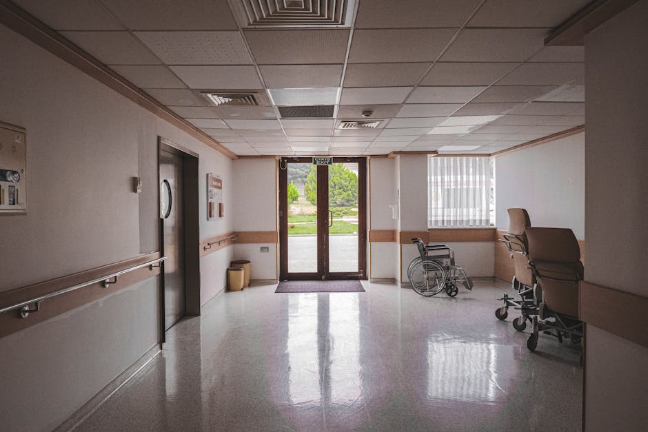 An image of a hospital corridor showing a clean, well-lit interior with a polished, reflective floor surface. To the right, there are two hospital beds with beige upholstery and metal frames, position