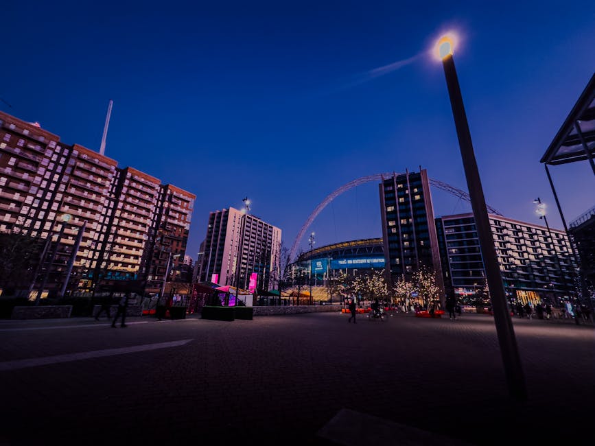 A cityscape scene during dusk featuring modern multi-storey buildings with illuminated windows, situated along a wide street with a few pedestrians walking on the pavement. In the background, a large Ferris wheel and a stadium are visible, lit with colorful lights. A tall streetlight stands prominently in the foreground, casting light onto the area. The environment appears calm with a clear evening sky, capturing the urban atmosphere that could be part of a home relocation or moving service area in Wembley as seen in the context of Man with Van Wembley’s removals services.