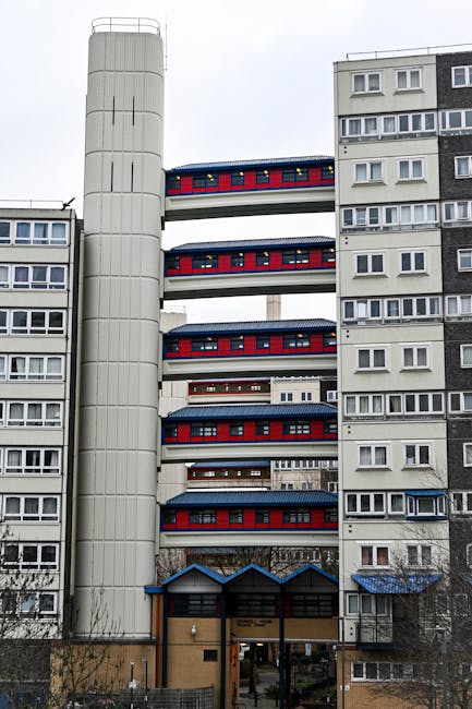 The exterior of a multi-storey residential building features a modern design with a prominent external elevator shaft on the left side, which is wrapped in a light grey casing. The building has several floors with visible balconies, each equipped with red and blue painted railings and dark roofing, indicating a possible renovation or decorative element. The balconies are open, and some have items such as small potted plants or furniture, suggesting residential use. The ground floor includes a covered entrance area with a small canopy, leading to the building’s lobby. Surrounding the building is a paved pavement and some sparse trees, indicating an urban setting. This image is relevant to home relocation and furniture transport services, illustrating the challenges or considerations of moving items into higher-floor flats, such as through stairwells or external lifts. Man with Van Wembley, specializing in removals, would manage logistics related to packing, loading, and transporting household belongings, especially in buildings like this with lift outages or accessibility issues during a house or flat relocation.