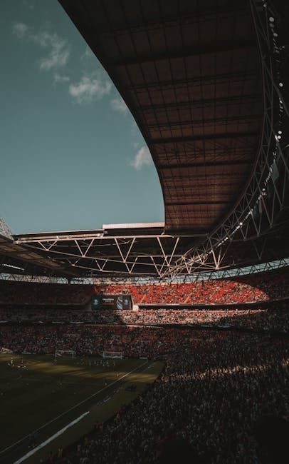 Interior view of a large football stadium with a well-maintained grass pitch, surrounded by multiple tiers of red seating in the stands. The stadium roof structure includes white steel trusses and a p
