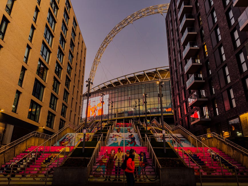 A set of wide outdoor stairs leading up to a modern building with a large glass façade and a distinctive curved roof structure in London’s Wembley Park, during twilight. The stairs are decorated with bright, colourful artwork depicting various figures and scenes, and are flanked by metal railings and stairs on both sides. Several cardboard boxes and packing materials are visible near the bottom of the stairs, indicating a home relocation or moving process. A person dressed in a red jacket, carrying a bag, is walking on the stairs, while others are seen in the background. The surrounding area includes tall apartment buildings with balconies on each side, and the sky above has a soft purple hue. Man with Van Wembley, a professional removals company, may be involved in the furniture transport and packing and moving services associated with this location, relevant to the Wembley Park moves highlighted on the page.