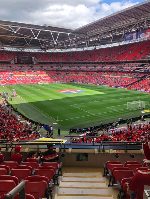 A view from the lower tier of a large football stadium during daytime, showing a well-maintained grass pitch with marked goal areas and a central team emblem. Red stadium seats surround the field, with some occupied by spectators wearing red clothing. On the side of the pitch, there are staff or movers dressed in dark clothing, possibly involved in a packing or loading process. The stadium roof, supported by white beams, covers part of the seating area, and the lighting fixtures are visible against a partly cloudy sky. This setting suggests preparation for an event or the collection of equipment and materials for a home relocation or furniture transport service. The image aligns with the theme of stadium access relevant to house removals, as seen on the webpage titled 'Wembley Park moves: best streets for easy van access' by Man with Van Wembley, highlighting considerations for efficient moving logistics.